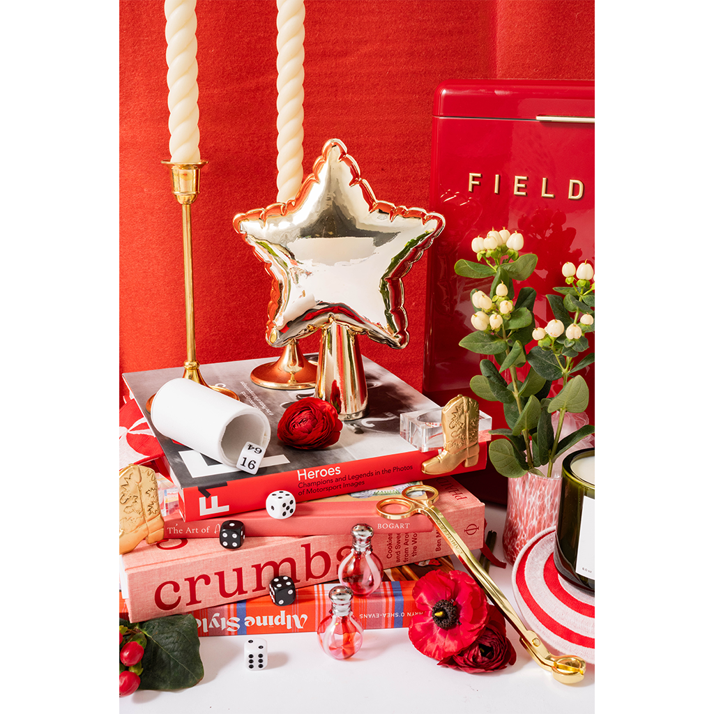 A styled tabletop arrangement featuring a metallic silver star-shaped balloon on a gold stand, red and pink books, white pillar candles, small perfume bottles, dice, flowers, and a red-and-white striped plate against a red backdrop.