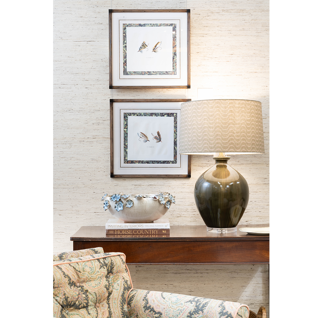 A polished interior vignette featuring a dark wood console table styled with a sculptural ceramic bowl, stacked books, and a glossy olive green table lamp with a patterned shade. Two framed bird illustrations hang above, while a patterned upholstered chair adds color and texture in the foreground.