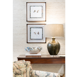 A polished interior vignette featuring a dark wood console table styled with a sculptural ceramic bowl, stacked books, and a glossy olive green table lamp with a patterned shade. Two framed bird illustrations hang above, while a patterned upholstered chair adds color and texture in the foreground.