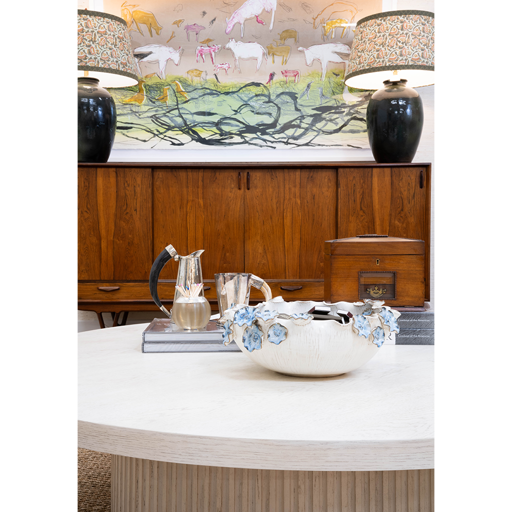 A layered interior featuring a large white cocktail table styled with a sculptural ceramic bowl, silver pitchers, books, and a wooden box. A warm wood cabinet is flanked by dark ceramic lamps with patterned shades, while colorful animal artwork creates a playful focal point above.