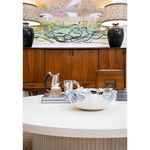 A layered interior featuring a large white cocktail table styled with a sculptural ceramic bowl, silver pitchers, books, and a wooden box. A warm wood cabinet is flanked by dark ceramic lamps with patterned shades, while colorful animal artwork creates a playful focal point above.
