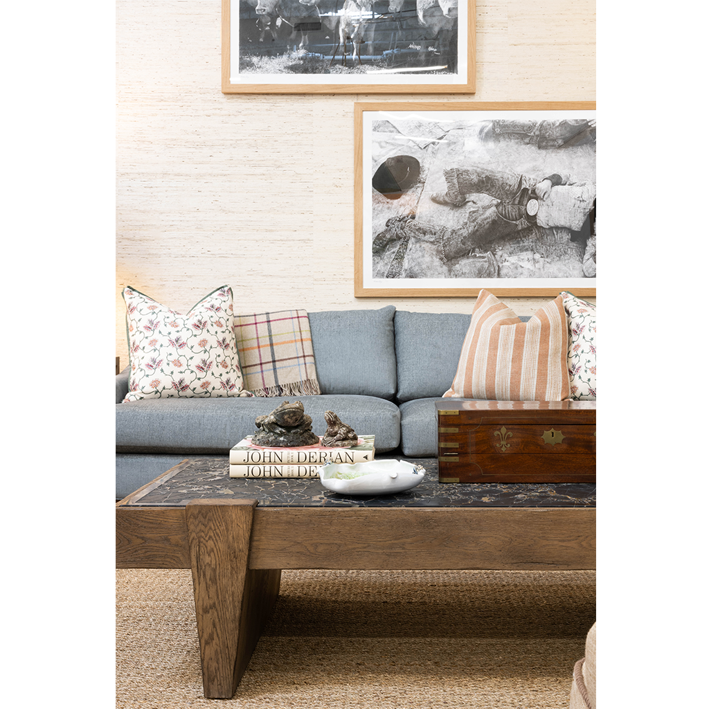 A cozy living room featuring a blue upholstered sofa layered with floral and striped pillows beneath framed black-and-white artwork. A substantial wood cocktail table with a dark stone top is styled with stacked books, decorative objects, and a vintage wooden box.