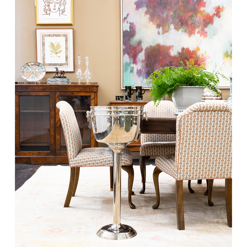 A sophisticated dining room featuring patterned upholstered chairs around a dark wood table, accented by a polished silver wine cooler on a pedestal stand. A large colorful abstract painting, lush green fern, framed artwork, and dark wood cabinet complete the layered interior.