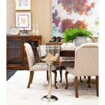 A sophisticated dining room featuring patterned upholstered chairs around a dark wood table, accented by a polished silver wine cooler on a pedestal stand. A large colorful abstract painting, lush green fern, framed artwork, and dark wood cabinet complete the layered interior.