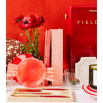 A styled tabletop scene with a red FIELD refrigerator, red flowers, a pink book, a pink glass bowl, a glass, and a wooden tray holding gold-toned utensils with red handles, set against a red wall.