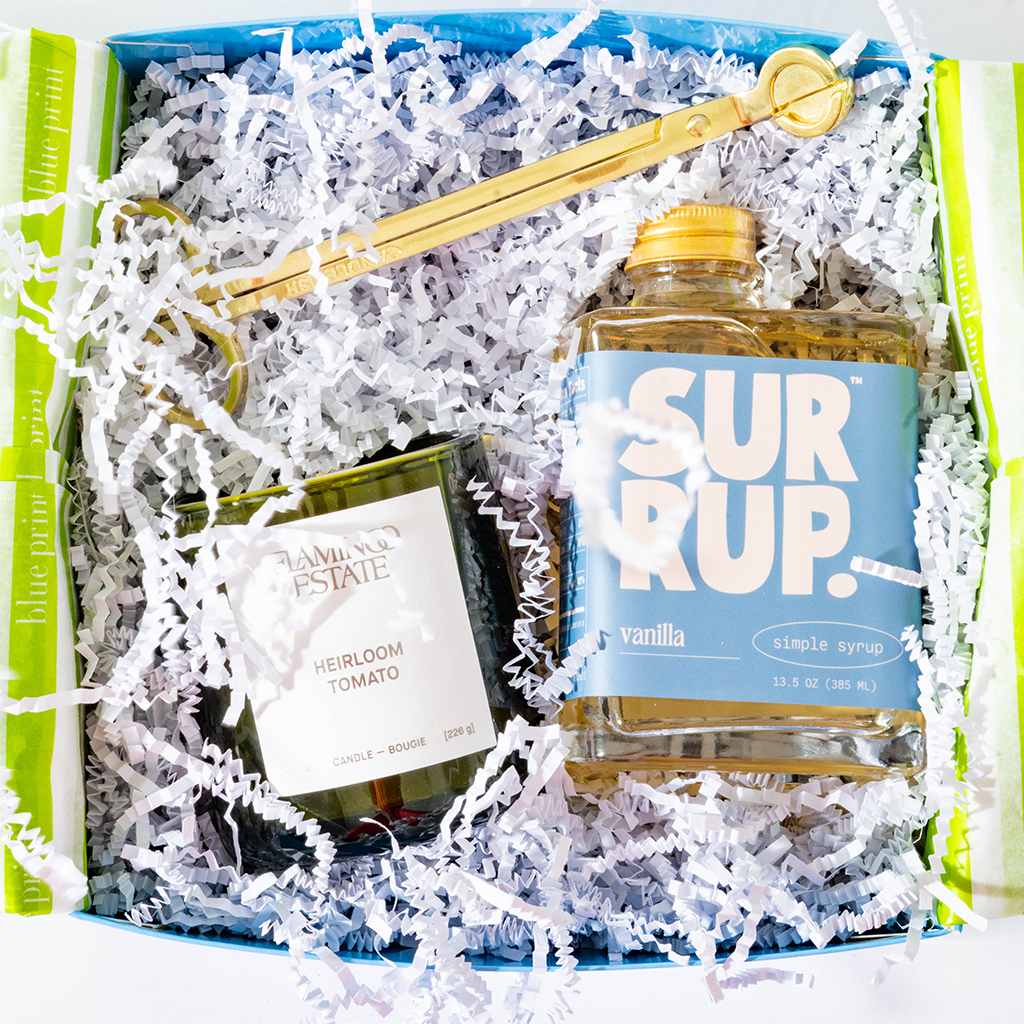 A gift box filled with white shredded paper containing a blue-labeled SURUP vanilla simple syrup bottle, a dark green candle jar labeled Hannah's Estate Heirloom Tomato, and a gold spoon resting across the top.