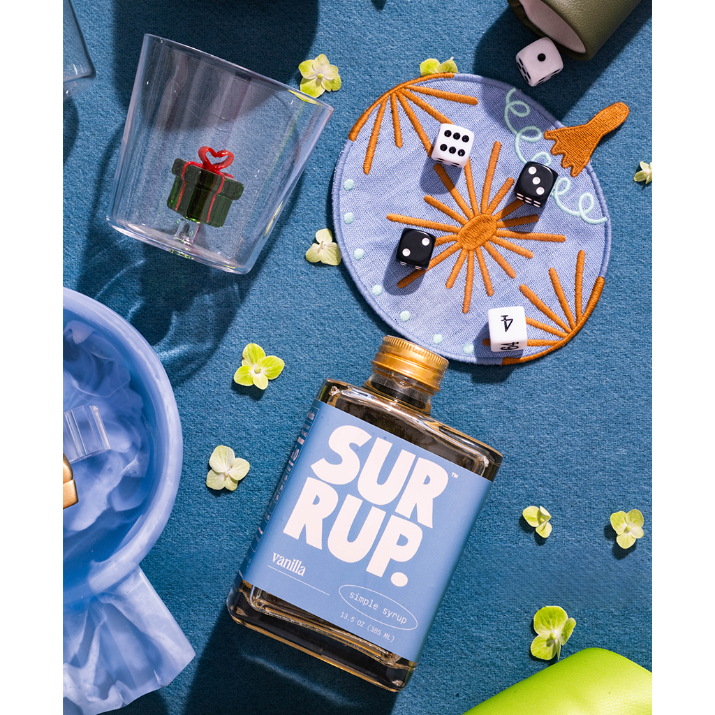 A bottle of SUR RUP. Vanilla flavored malt beverage with a blue label and gold cap, styled on a blue textured surface with a clear gift-themed tumbler, embroidered plate, dice, and small yellow flowers.