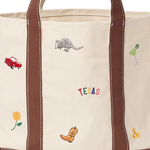 a close up of An off-white canvas tote bag with brown leather handles and base, decorated with small embroidered western-themed motifs including a cowboy boot, an armadillo, a colorful Texas sign, flowers, and a margarita