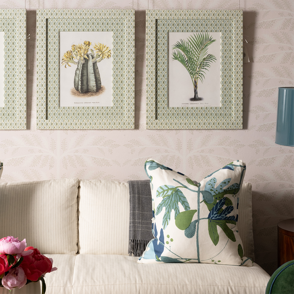 Decorative pillow with green and blue botanical leaf print on beige sofa under framed palm and floral artwork.
