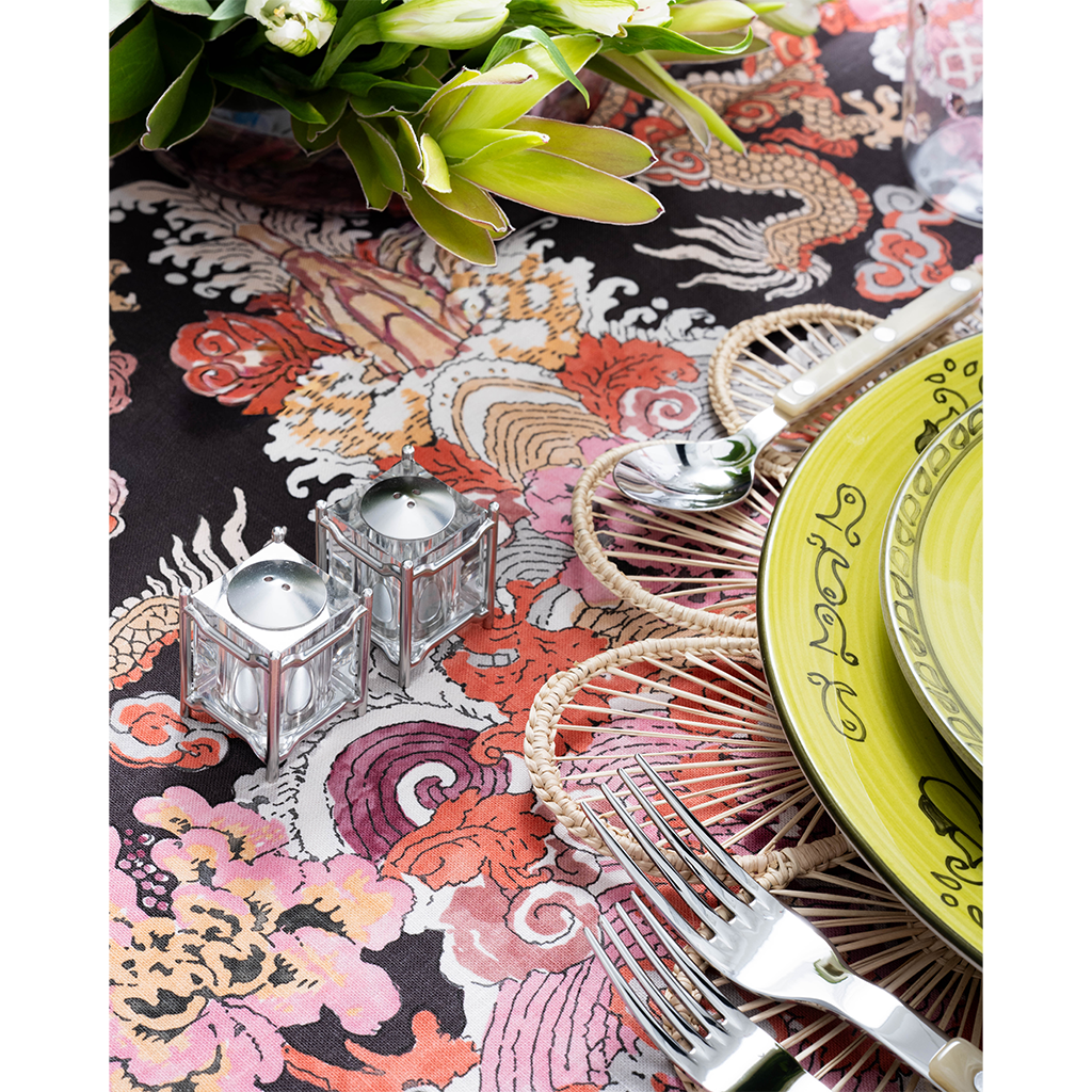 A close-up of a vibrant tablescape featuring a bold multicolored dragon-print tablecloth, woven floral placemat, lime green patterned plates, and pearl-handled flatware. Two small glass candle holders and a lush green floral arrangement add decorative detail to the setting.
