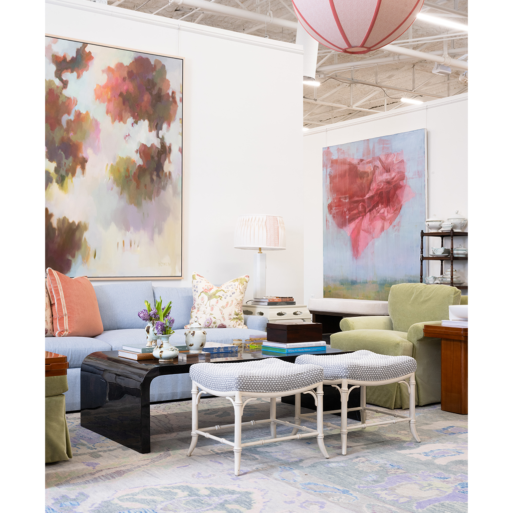 A styled living room interior featuring two large abstract wall art prints hanging above a light blue sofa. The room includes a black coffee table, two white upholstered stools, a green armchair, a lamp, and a patterned area rug.