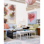 A styled living room interior featuring two large abstract wall art prints hanging above a light blue sofa. The room includes a black coffee table, two white upholstered stools, a green armchair, a lamp, and a patterned area rug.