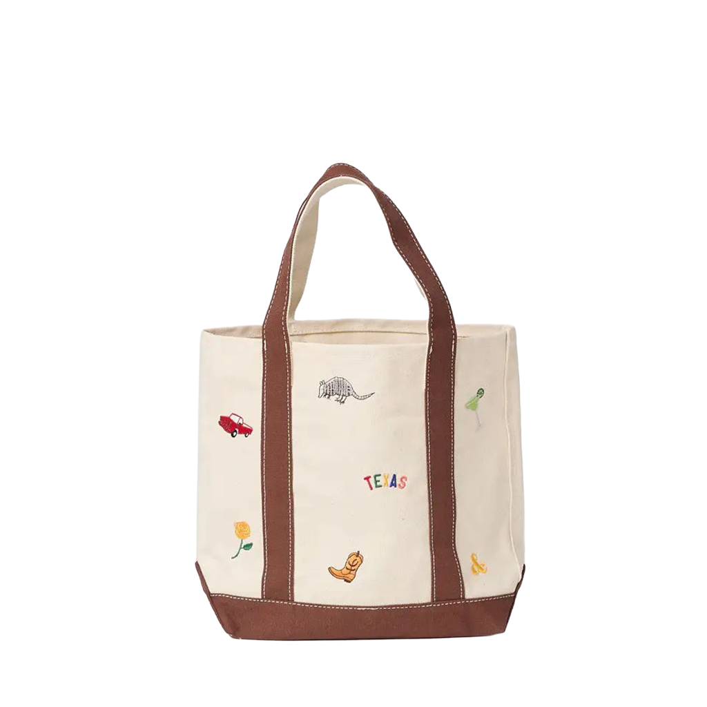 An off-white canvas tote bag with brown leather handles and base, decorated with small embroidered western-themed motifs including a cowboy boot, an armadillo, a colorful Texas sign, flowers, and a margarita