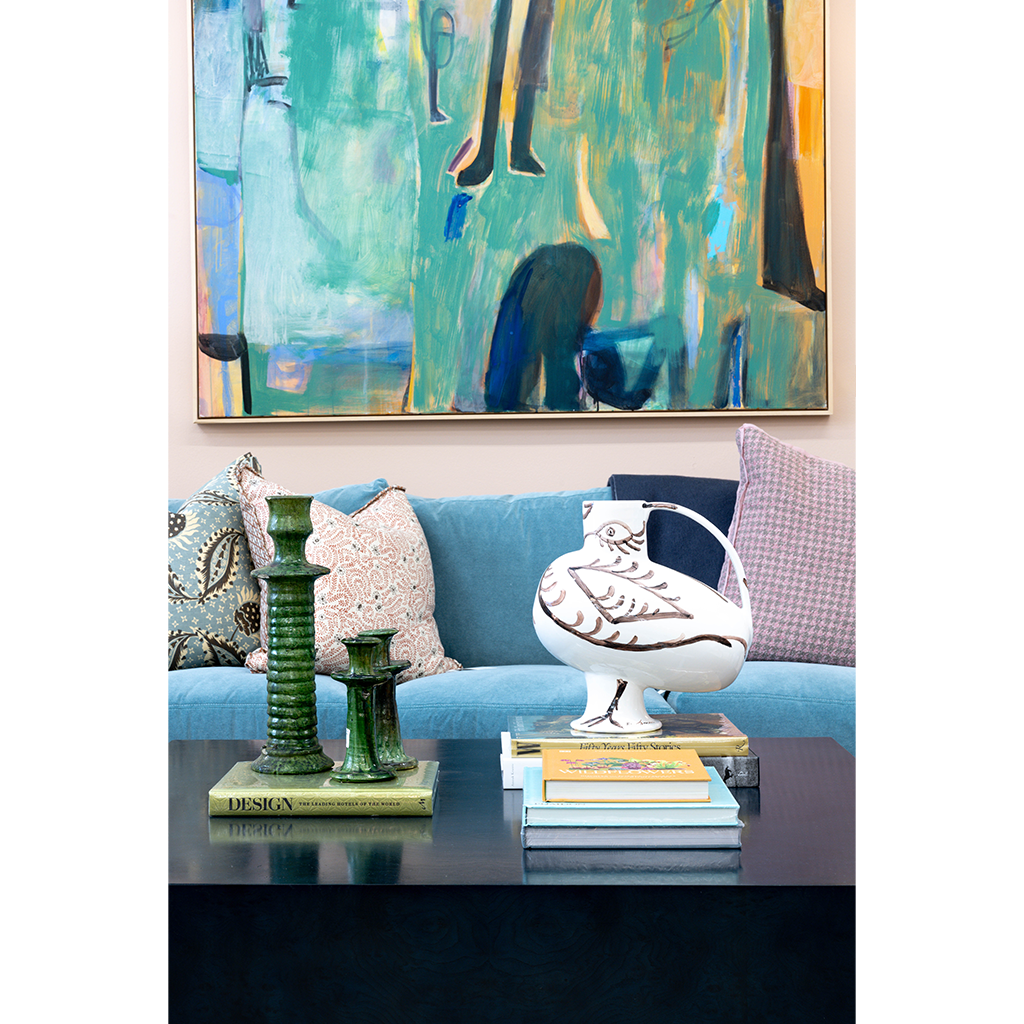 Sculptural white ceramic bird vessel with hand-painted brown details displayed on stacked books atop a dark cocktail table. Green glass candlesticks, colorful patterned pillows, a bright blue sofa, and large abstract artwork create an eclectic, layered setting around the piece.