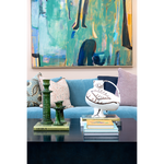 Sculptural white ceramic bird vessel with hand-painted brown details displayed on stacked books atop a dark cocktail table. Green glass candlesticks, colorful patterned pillows, a bright blue sofa, and large abstract artwork create an eclectic, layered setting around the piece.