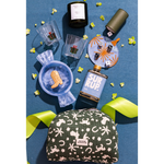 A styled flat-lay of a holiday-themed gift set on a textured blue background, featuring a green and pink bunny print pouch, a blue candy cane candle, a 'SUR RUP.' bottle, and other festive accessories.