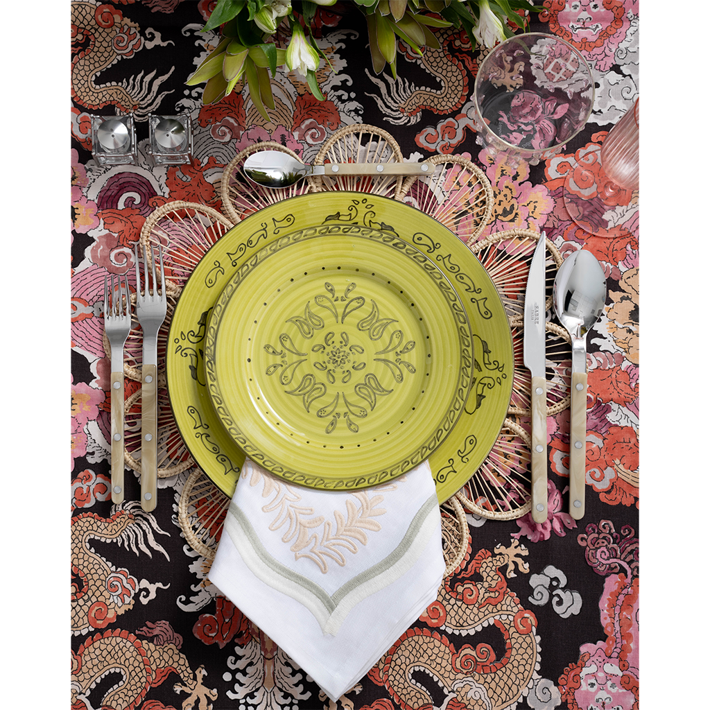 A vibrant place setting featuring a lime green patterned plate layered over a woven floral-shaped placemat and embroidered white napkin. Pearl-handled flatware, pink glassware, and a bold multicolored dragon-print tablecloth complete the playful tablescape.