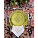 A vibrant place setting featuring a lime green patterned plate layered over a woven floral-shaped placemat and embroidered white napkin. Pearl-handled flatware, pink glassware, and a bold multicolored dragon-print tablecloth complete the playful tablescape.