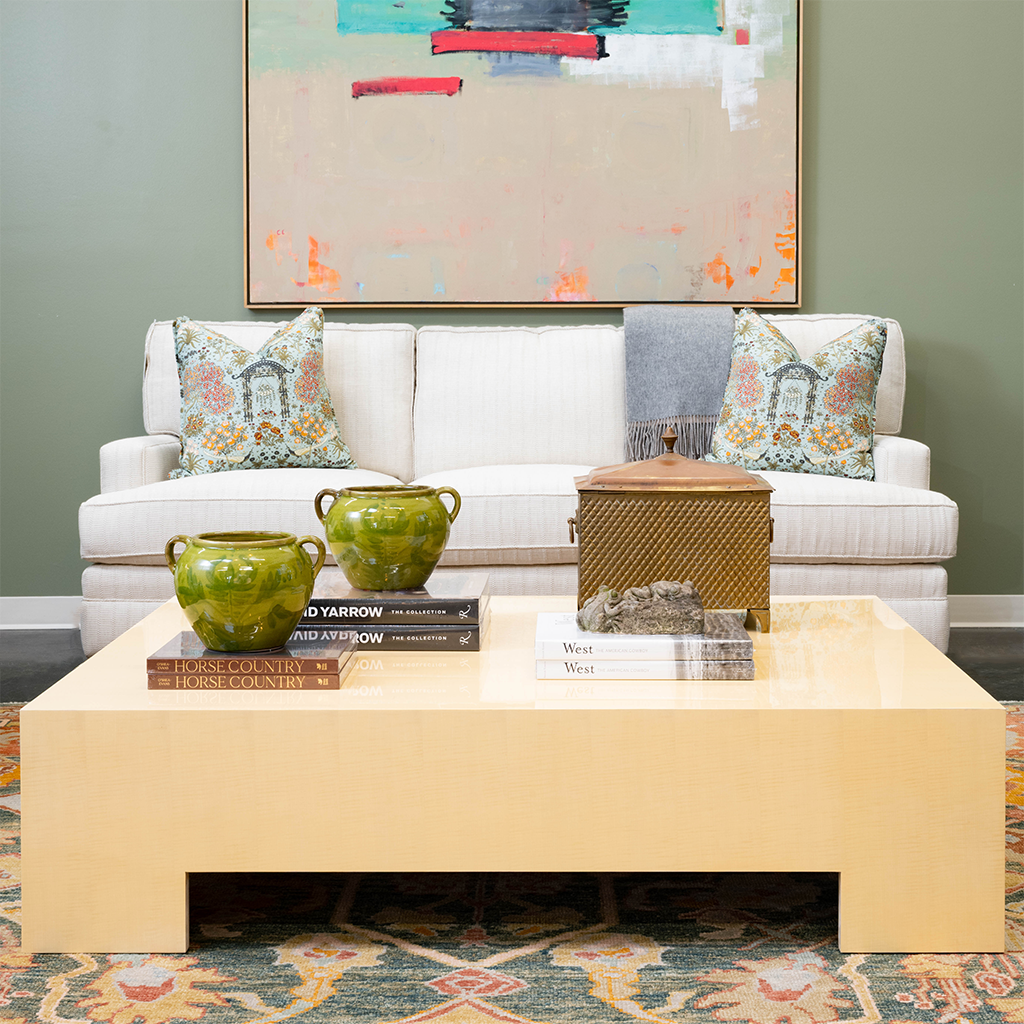 Bright living room featuring a large cream lacquered cocktail table styled with green ceramic vessels, decorative books, and a woven box. A white upholstered sofa with patterned pillows sits beneath colorful abstract artwork against soft green walls.