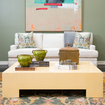Bright living room featuring a large cream lacquered cocktail table styled with green ceramic vessels, decorative books, and a woven box. A white upholstered sofa with patterned pillows sits beneath colorful abstract artwork against soft green walls.