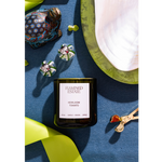 A Flamingo Estate 'Herbloom Tomato' scented candle in a dark glass jar, styled in a flat-lay arrangement on a textured blue surface with decorative green leaves, a patterned figurine, and a white fabric piece.