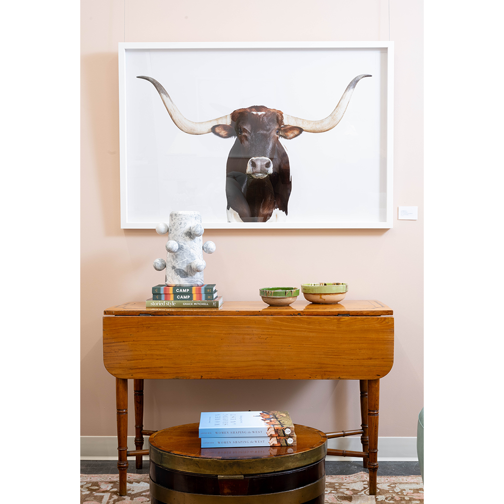 A warm interior vignette featuring a wooden drop-leaf console table styled with decorative bowls, stacked books, and a sculptural accent. A large framed longhorn portrait hangs above, while a round wood and brass coffee table with books sits in the foreground.