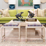 A colorful living room featuring a lime green upholstered sofa with patterned throw pillows and a pair of blue ceramic lamps. Two plaid-cushioned stools sit in front of a light wood cocktail table styled with decorative horse sculptures, a ceramic bowl, and a patterned box.
