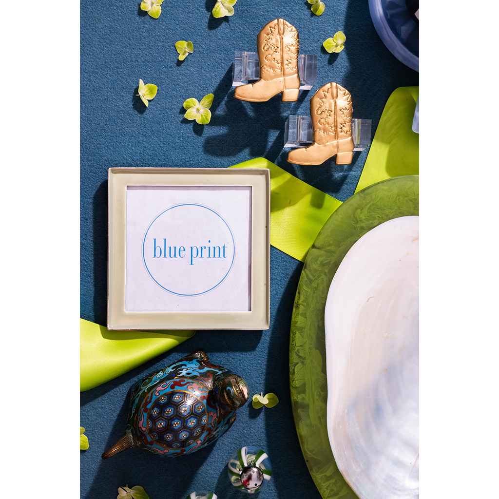 A framed print with the words 'blue print' in blue text on a white background, styled in a flat lay with small cowboy boot-shaped clips, green ribbon, a decorative peacock figurine, and a white plate on a dark blue surface.