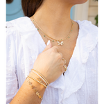 A close-up lifestyle shot of a woman wearing a layered gold necklace with a butterfly pendant and green gemstone accents, along with matching gold bracelets and rings, paired with a white ruffled blouse.