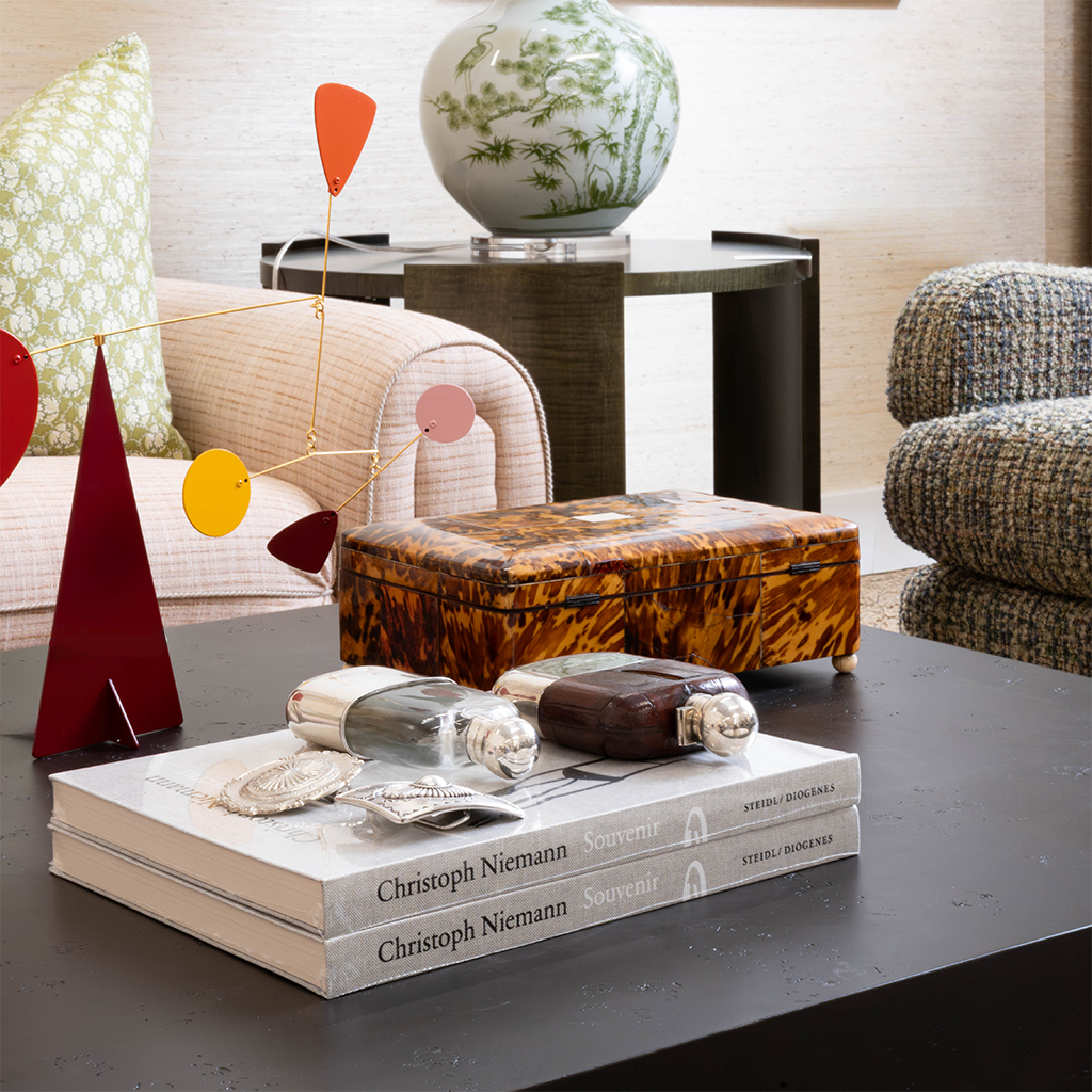 A layered living room vignette featuring a dark cocktail table styled with stacked art books, decorative bottles, a tortoiseshell box, and a colorful sculptural mobile. A green-and-white ceramic vase, blush upholstered chair, and textured seating add pattern and color to the space.