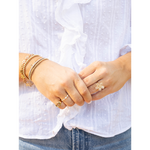 A close-up of a person's hands wearing multiple gold-toned jewelry pieces, including bracelets, rings, and a flower-shaped ring, styled with a white eyelet blouse and blue jeans.