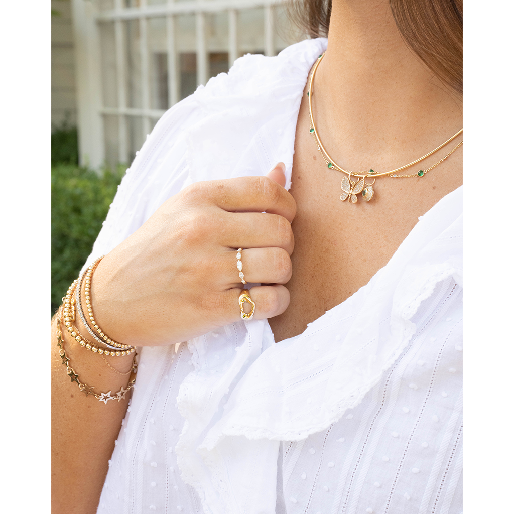 A close-up lifestyle shot of a woman wearing delicate gold jewelry, including a gold necklace with a butterfly and green bead accents, a gold ring, and layered gold bracelets, paired with a white blouse.