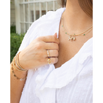 A close-up lifestyle shot of a woman wearing delicate gold jewelry, including a gold necklace with a butterfly and green bead accents, a gold ring, and layered gold bracelets, paired with a white blouse.