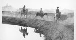 Three cowboys on horseback near a water hole with reflections in the water.