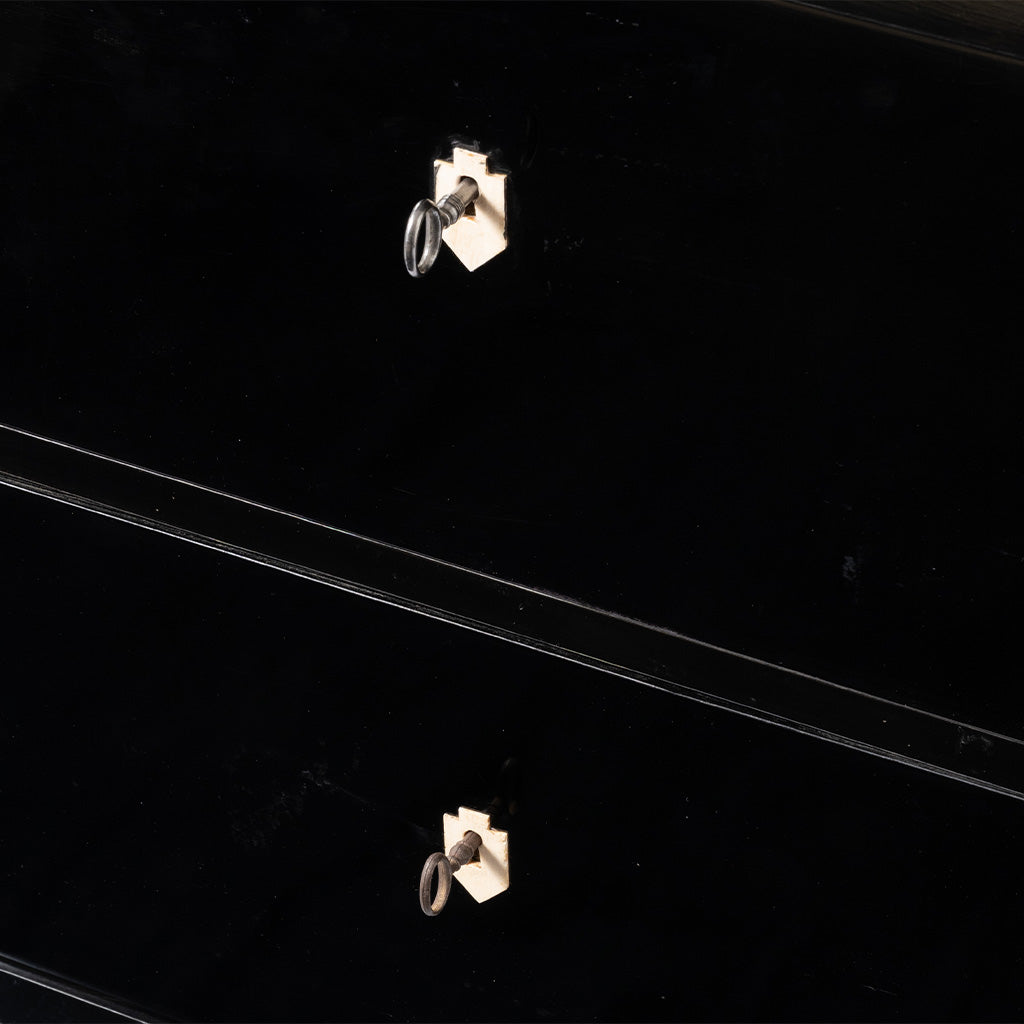 A close-up of an antique ebonized chest of drawers highlights the glossy black finish and ivory-colored shield-shaped keyhole escutcheons, each fitted with its original key. The image emphasizes the chest's clean lines and understated craftsmanship.