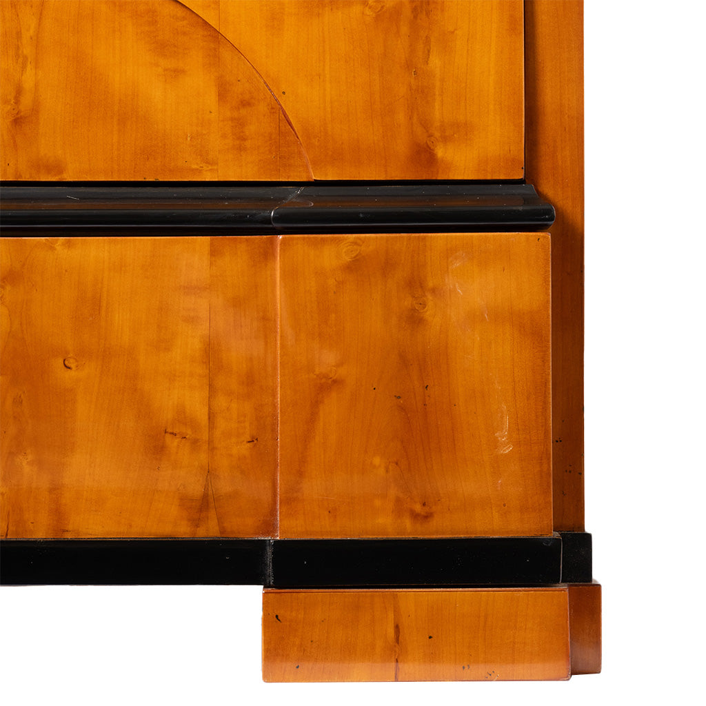 A close-up of an antique Biedermeier chest of drawers highlights the warm honey-colored wood veneer, contrasting ebonized trim, and stepped block foot. The image emphasizes the clean lines and fine craftsmanship characteristic of the Biedermeier style.