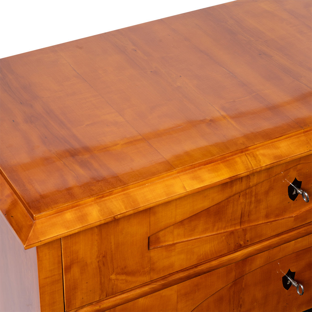 A close-up of an antique Biedermeier chest of drawers highlights the polished honey-colored wood top with beautifully figured veneer and a beveled edge. The image also shows the upper drawer fronts with geometric veneer detailing and traditional keyhole escutcheons.