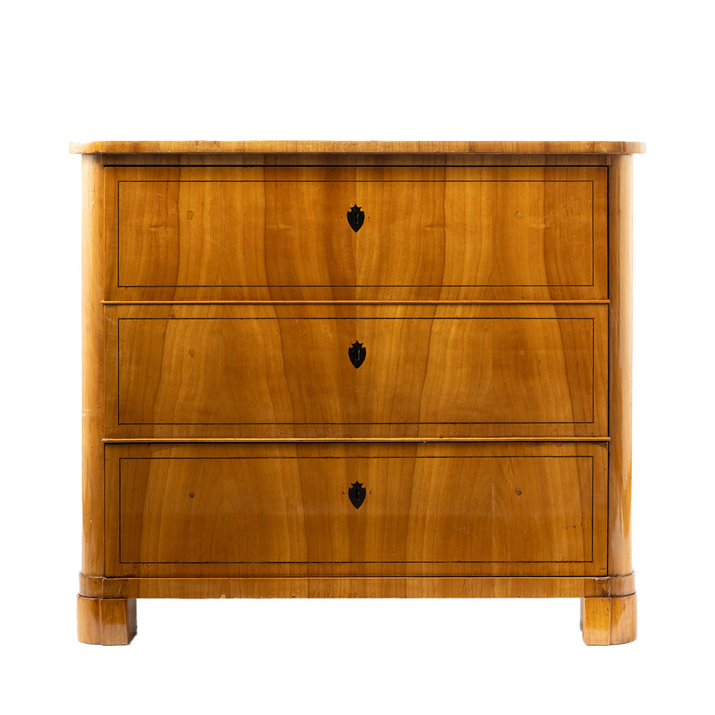 A front facing view of an antique Biedermeier chest of drawers in a warm honey-colored wood veneer features three graduated drawers with keyholes and delicate string inlay. Rounded front corners, clean lines, and block feet emphasize the piece's refined, understated design.