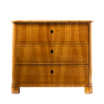 A front facing view of an antique Biedermeier chest of drawers in a warm honey-colored wood veneer features three graduated drawers with keyholes and delicate string inlay. Rounded front corners, clean lines, and block feet emphasize the piece's refined, understated design.