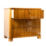 A three-quarter view of an antique chest of drawers with a warm honey-colored wood veneer features three graduated drawers with keyholes and subtle string inlay. The clean lines, rounded front corners, and block feet reflect the elegant simplicity of the Biedermeier style.