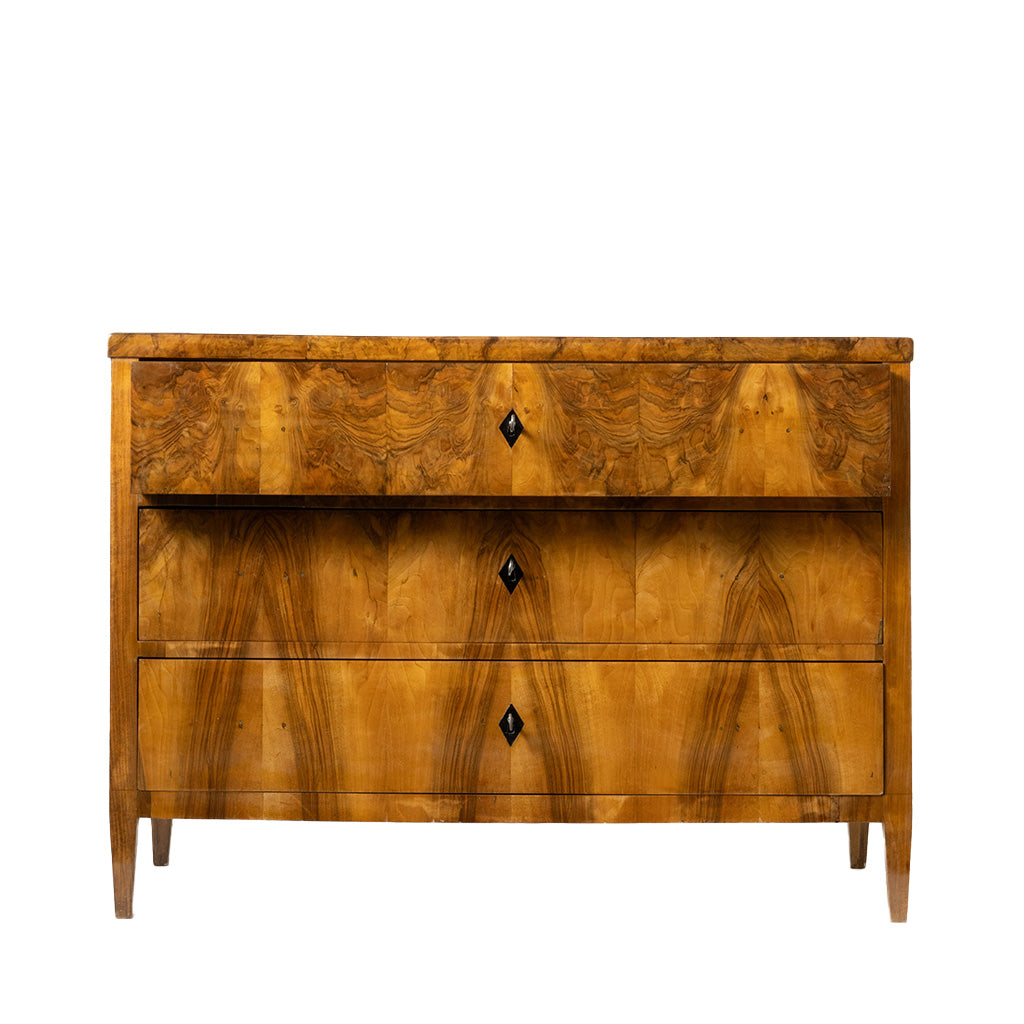 A front facing view of an antique Biedermeier chest of drawers shows the top drawer partially open, revealing dovetail joinery and a solid wood interior. The richly figured walnut veneer, dramatic bookmatched grain, and simple black diamond-shaped keyhole escutcheons highlight the piece's fine craftsmanship.