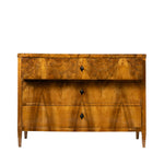 A front facing view of an antique Biedermeier chest of drawers shows the top drawer partially open, revealing dovetail joinery and a solid wood interior. The richly figured walnut veneer, dramatic bookmatched grain, and simple black diamond-shaped keyhole escutcheons highlight the piece's fine craftsmanship.