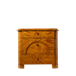 A front view of an antique Biedermeier chest of drawers revealing dovetail joinery and a solid wood interior. The richly figured golden burl wood veneer, arched veneer detailing across the lower drawers, and block feet highlight the piece's exceptional craftsmanship and elegant design.