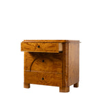 A three-quarter view of an antique Biedermeier chest of drawers shows the top drawer partially open, revealing dovetail joinery and a solid wood interior. The richly figured golden burl wood veneer, arched veneer detailing across the lower drawers, and block feet highlight the piece's exceptional craftsmanship and elegant design.