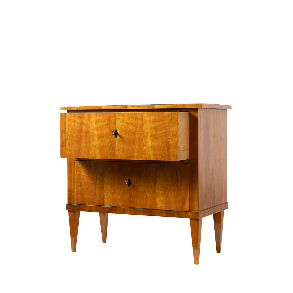 A three-quarter view of an antique Biedermeier two-drawer chest shows the top drawer partially open, revealing dovetail joinery and a solid wood interior. The warm honey-colored wood veneer, beautifully matched figured grain, tapered legs, and simple diamond-shaped pulls highlight the piece's refined craftsmanship.
