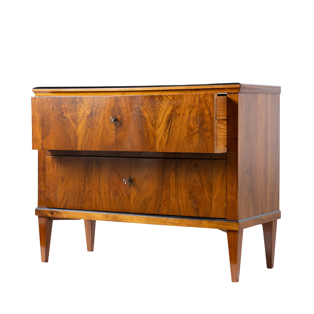 A three-quarter view of an antique Biedermeier two-drawer chest shows the top drawer partially open, revealing dovetail joinery and a solid wood interior. The striking bookmatched walnut veneer, tapered legs, and clean lines highlight the piece's exceptional craftsmanship and elegant design.