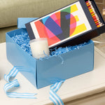 White candle with wooden wick in blue gift box filled with blue shredded paper and colorful abstract lid design.