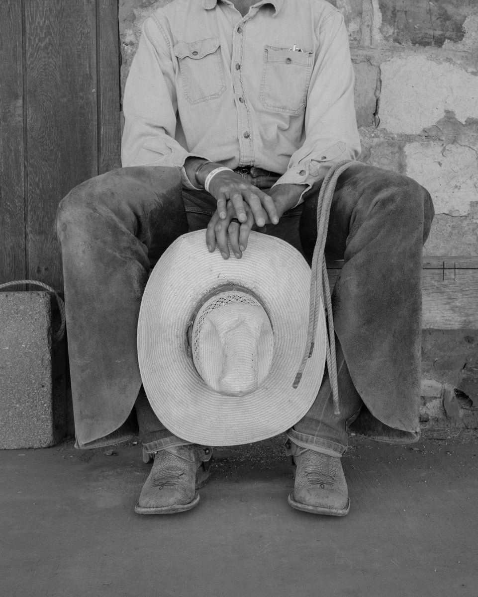 A black and white photograph of a person sitting on a rustic wooden bench, wearing a light button-up shirt, dark pants, boots, and holding a wide-brimmed cowboy hat in front of their lap.