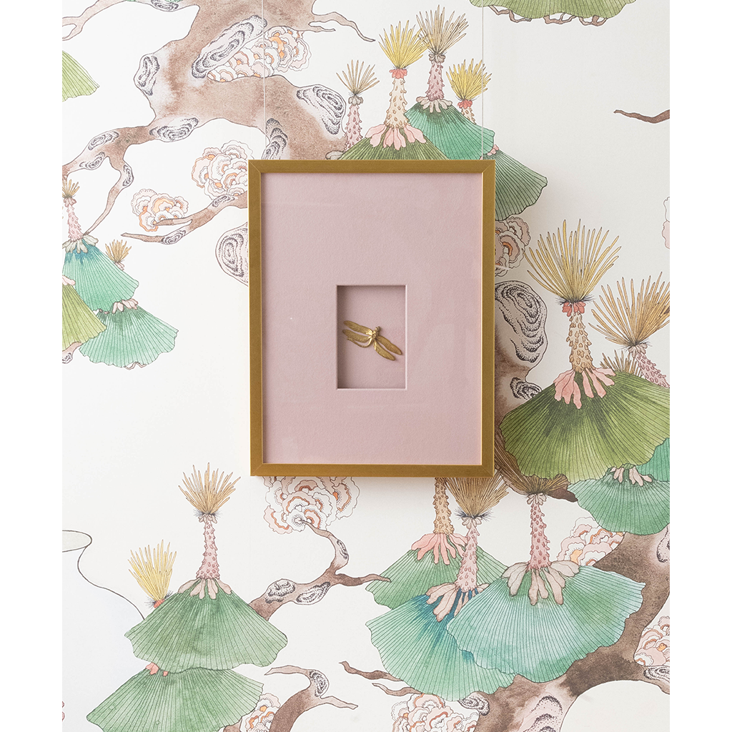 A square framed art piece with a light pink mat and a dragonfly in the center, mounted on a white wallpaper with a botanical pattern.