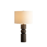 A modern table lamp with a dark brown wood-grain base featuring stacked cylindrical and spherical elements, topped with a warm off-white fabric drum shade.